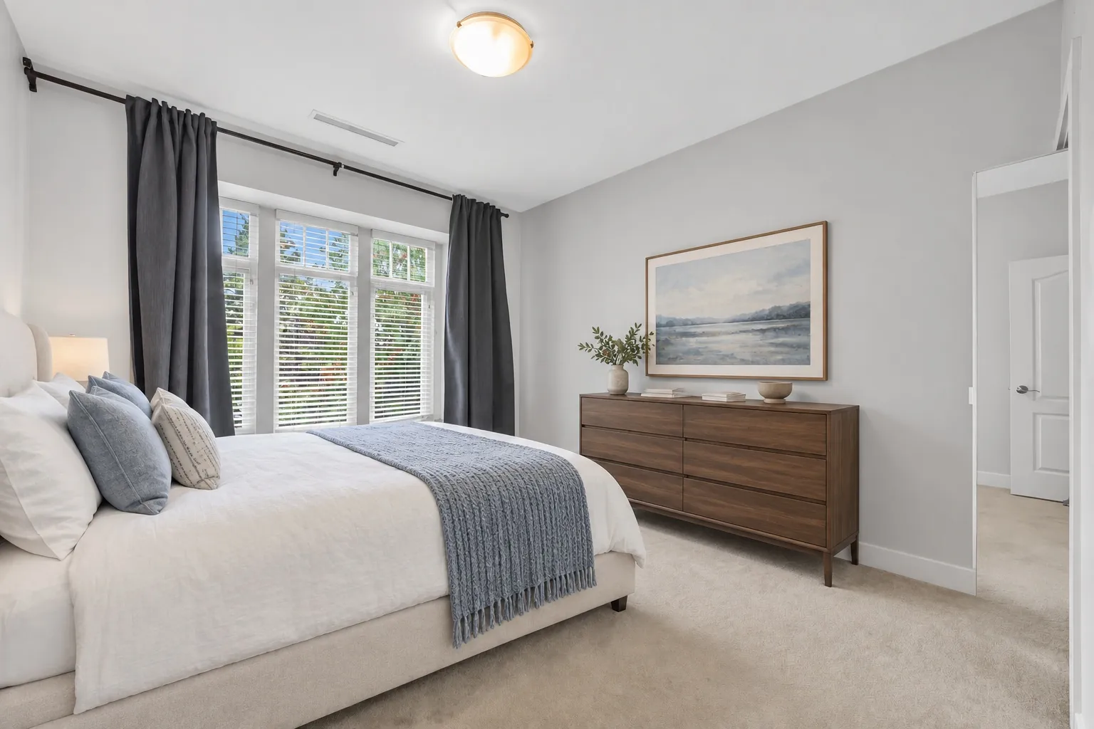 A bedroom virtually staged with a dressed bed, a wooden dresser and a picture, the window with grey curtains