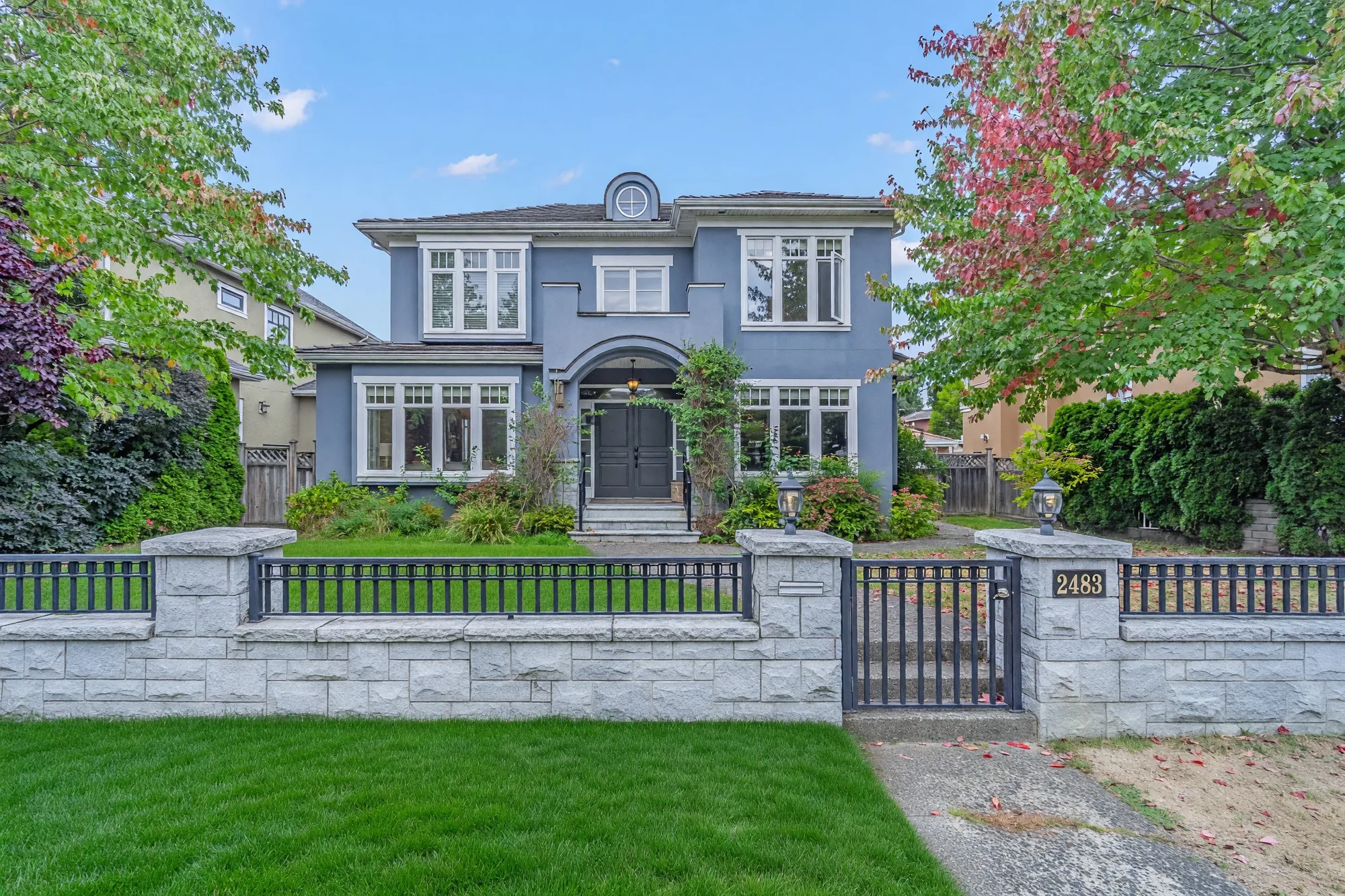 The front of the house in daylight: grey stucco, white-trimmed windows, the arched entry, a granite wall with an iron fence and the gate with the number 2483, a maple turning red at right