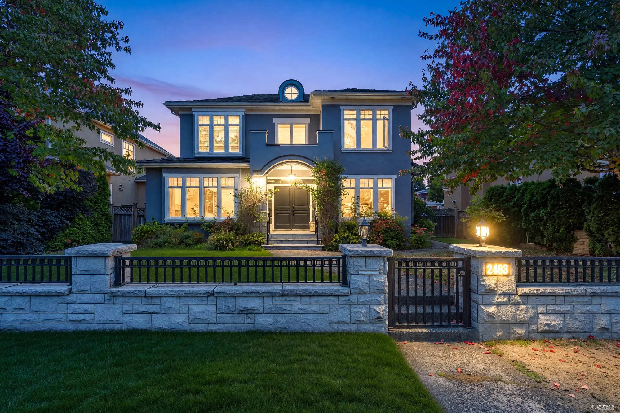 2483 West 22nd Avenue from the street at dusk: a grey stucco house with a hipped roof, its windows lit, behind a granite wall and iron fence with a lamp on the gate pillar