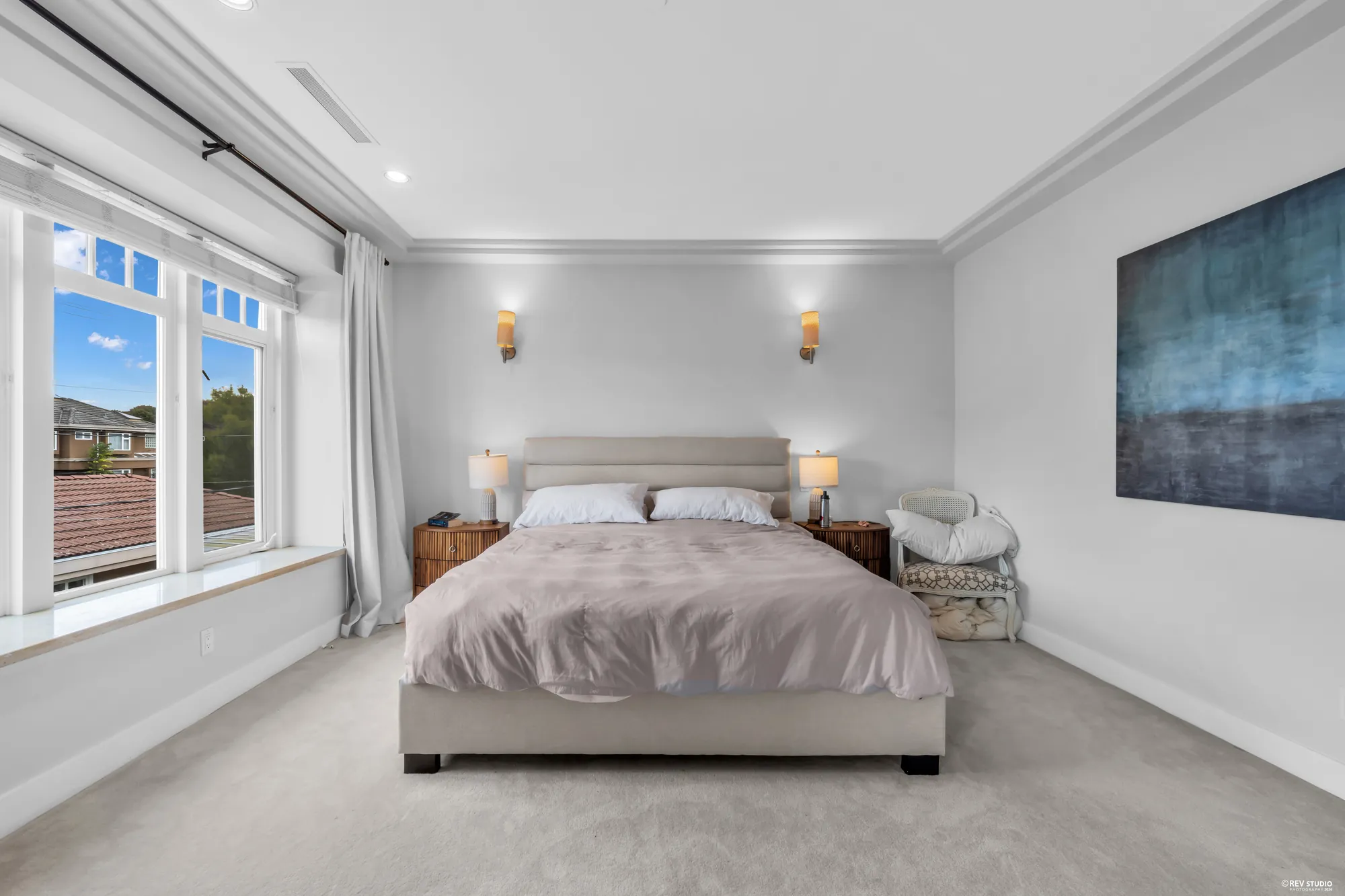 The primary bedroom: the bed between two sconces, windows on two walls, a large painting