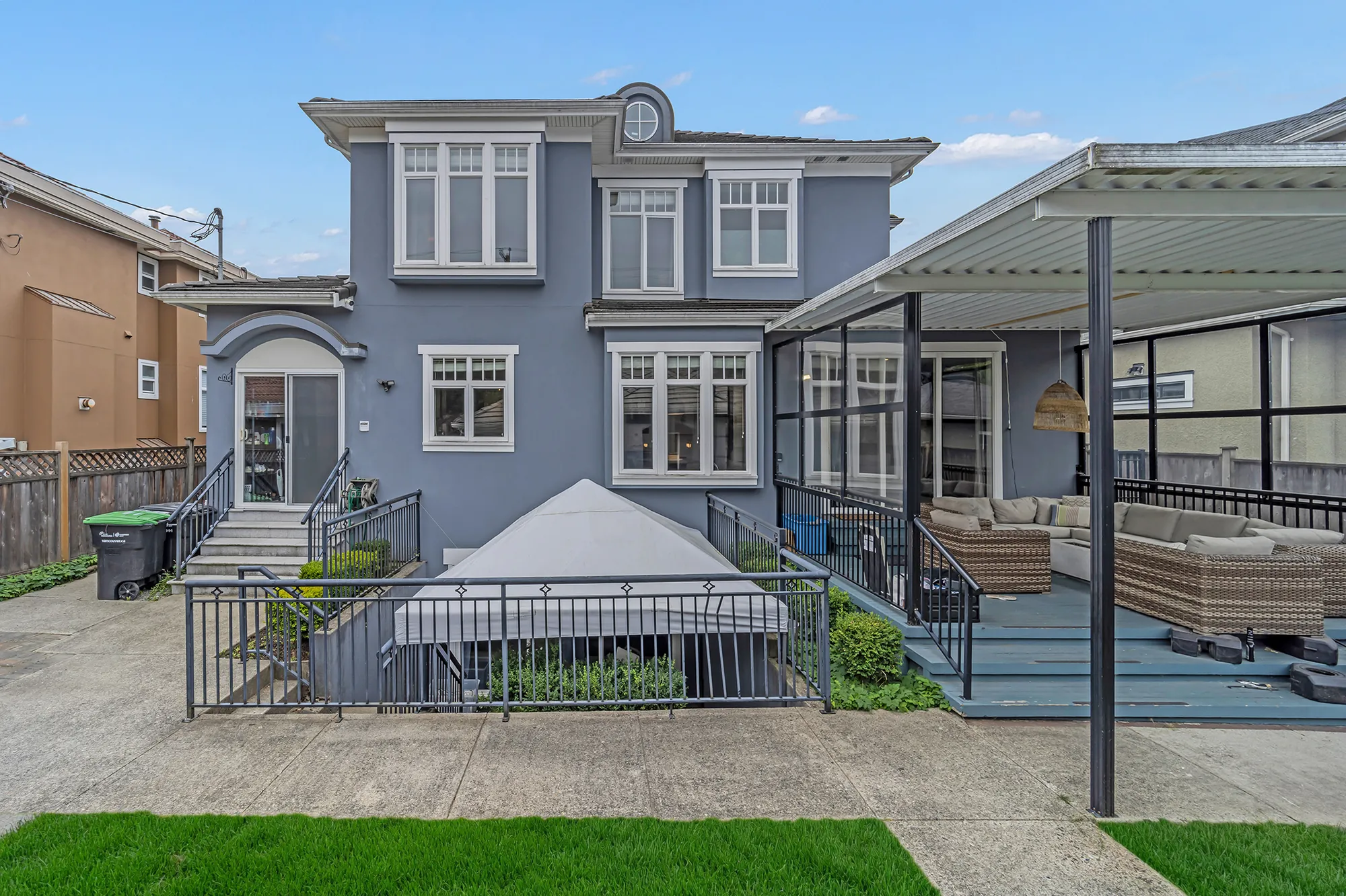 The back of the house in daylight: the covered deck with its outdoor sofas, the stair down to the canopied patio, the back door