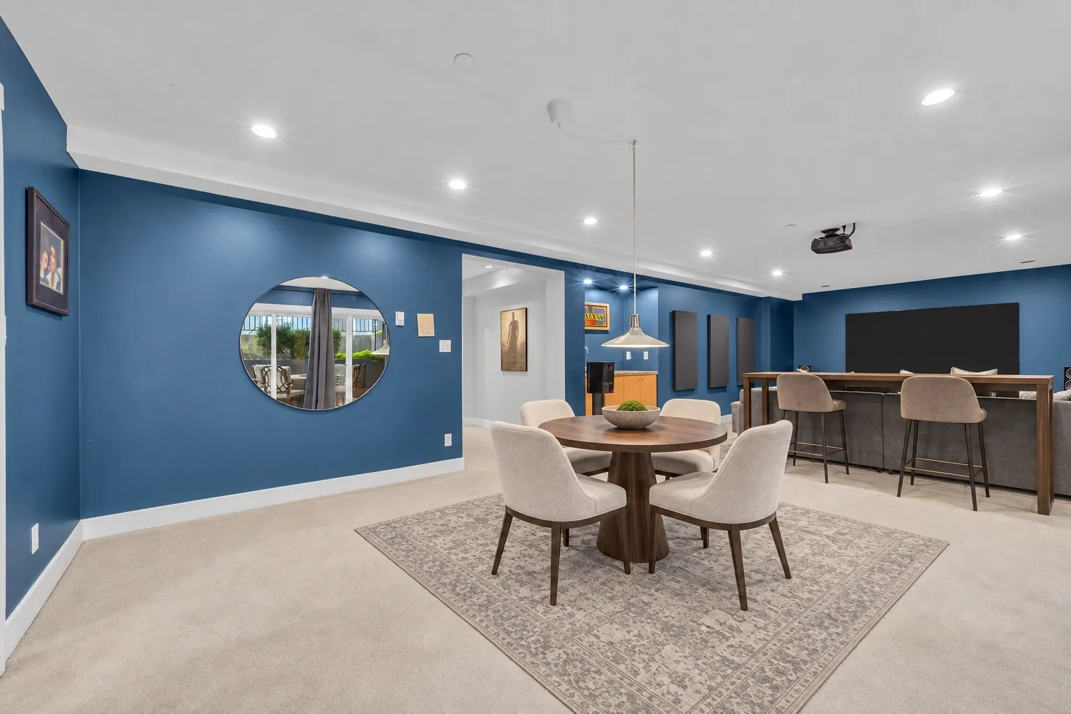 The recreation room toward the theatre room, a round mirror on the navy wall, virtually staged with a table and chairs and a bar with stools